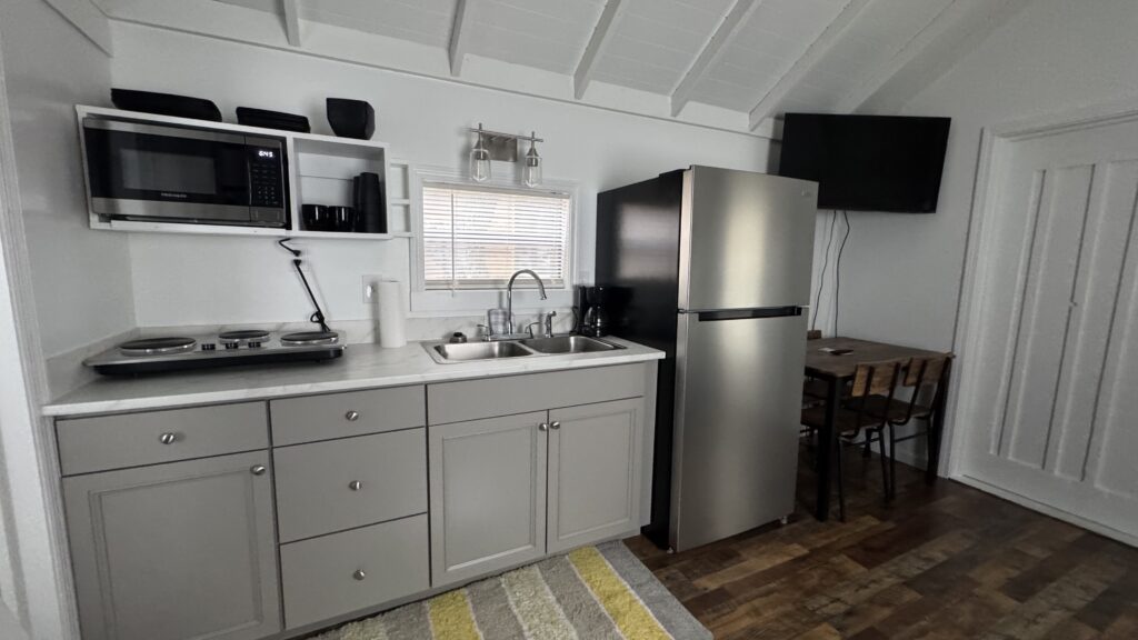Compact kitchen with gray cabinets, a sink, a microwave, a stovetop, a refrigerator, a wall-mounted TV, and a small dining table with two chairs on wooden flooring.