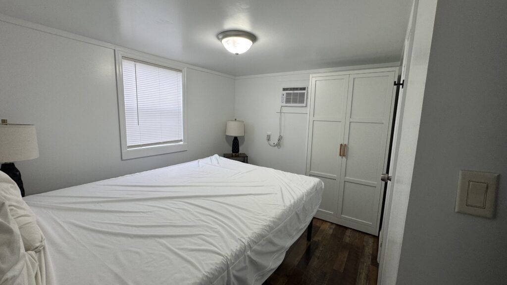 Small bedroom with a made bed, two lamps, a window with blinds, a wall-mounted air conditioner, and a white wardrobe against white walls and a dark wood floor.