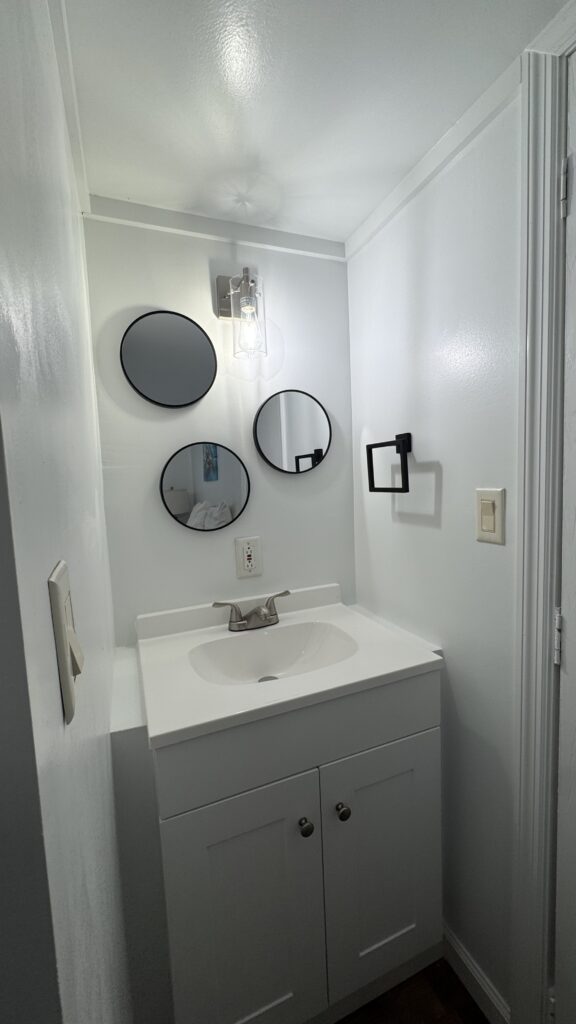 Small white bathroom with a modern vanity, three round mirrors, wall light fixture, towel ring, and light switch on a plain white wall.