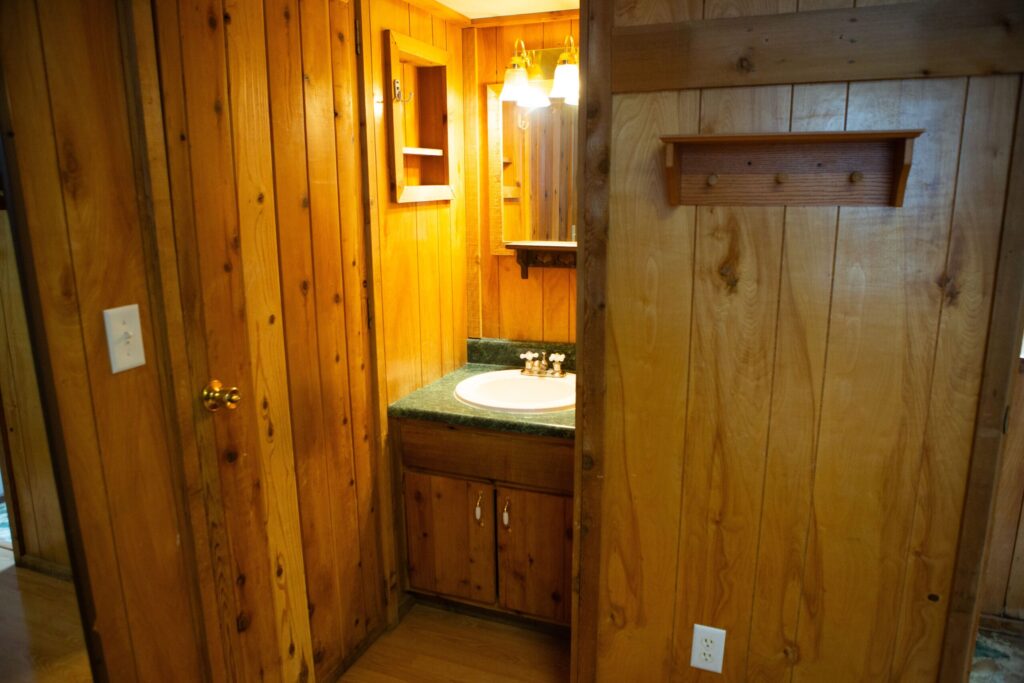 Small bathroom vanity area with a sink, green countertop, wooden cabinets, mirror, and wall-mounted lights, surrounded by wood-paneled walls.