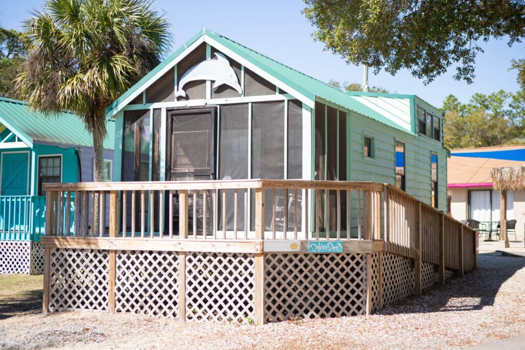 A small green cabin with a screened porch, wooden deck, and ramp, surrounded by palm trees and neighboring colorful buildings.