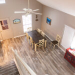 Overhead view of a dining room with a long dark table, eight white chairs, wood-look flooring, a ceiling fan, sideboard with lamps, and artwork on light-colored walls.