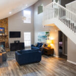 A modern living room with blue sofas, wooden furniture, a stone fireplace, TV, lamps, artwork, and a staircase leading to an upper floor.