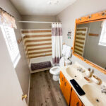 A bathroom with a bathtub and shower, toilet, double sink vanity with a large mirror, towels, and wood-style flooring. Light enters from a window with blinds and a valance.