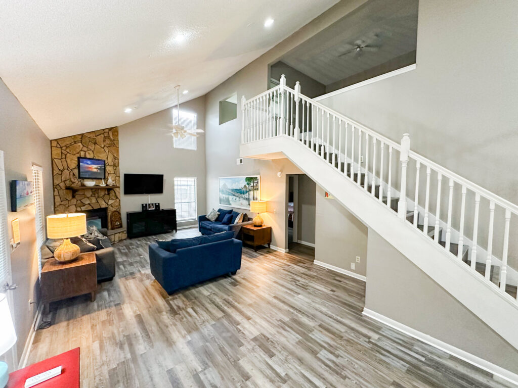 Spacious living room with vaulted ceiling, stone fireplace, blue sofas, TV, large windows, and white staircase leading to an upper floor.