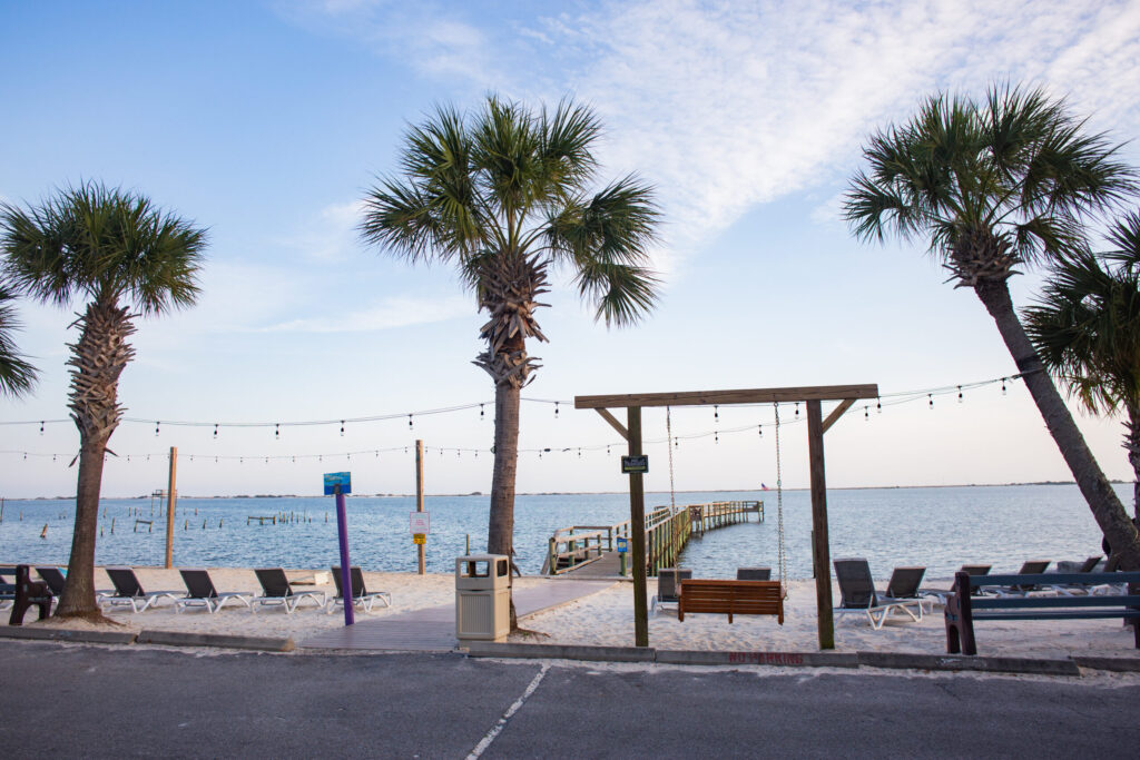 Palm trees line a sandy beach area with lounge chairs, a wooden dock extending into the water, string lights, and a partly cloudy sky.