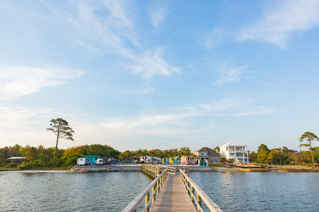 A wooden pier extends over the water toward a shoreline with houses, trees, and parked vehicles under a partly cloudy sky.