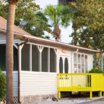 Single-story house with a screened porch, white siding, and a bright yellow wheelchair ramp, surrounded by palm trees and bushes.