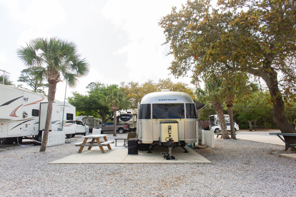 A silver Airstream trailer is parked on a gravel lot next to a picnic table, surrounded by trees and other RVs at a campground.