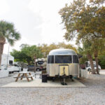 A silver Airstream trailer is parked on a gravel lot next to a picnic table, surrounded by trees and other RVs at a campground.