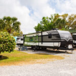 Several RVs are parked on gravel at a campground, with a small tree, picnic table, and palm trees visible under a partly cloudy sky.