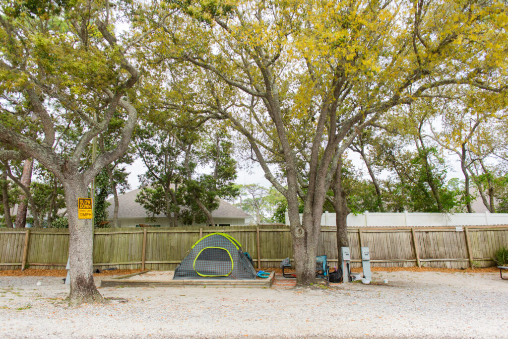 A green and gray tent is set up under large trees in a gravel campsite, with camping gear nearby and a wooden fence and houses in the background.