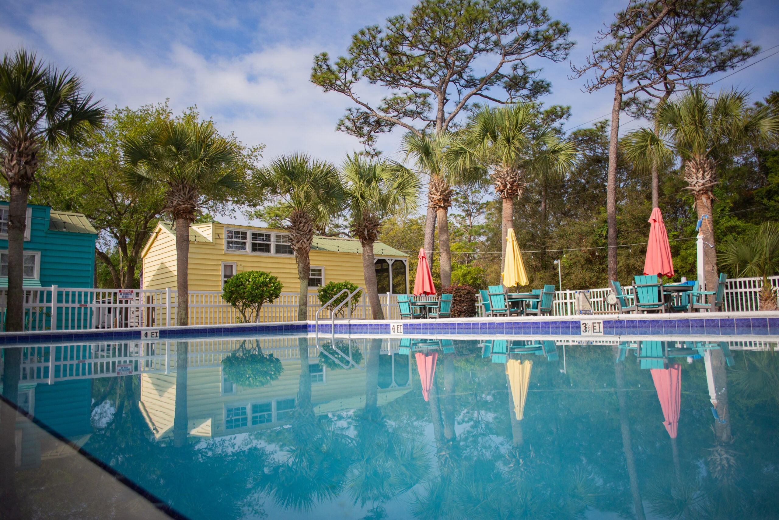 Outdoor swimming pool with reflections of palm trees, colorful patio umbrellas, and small buildings in the background under a partly cloudy sky.