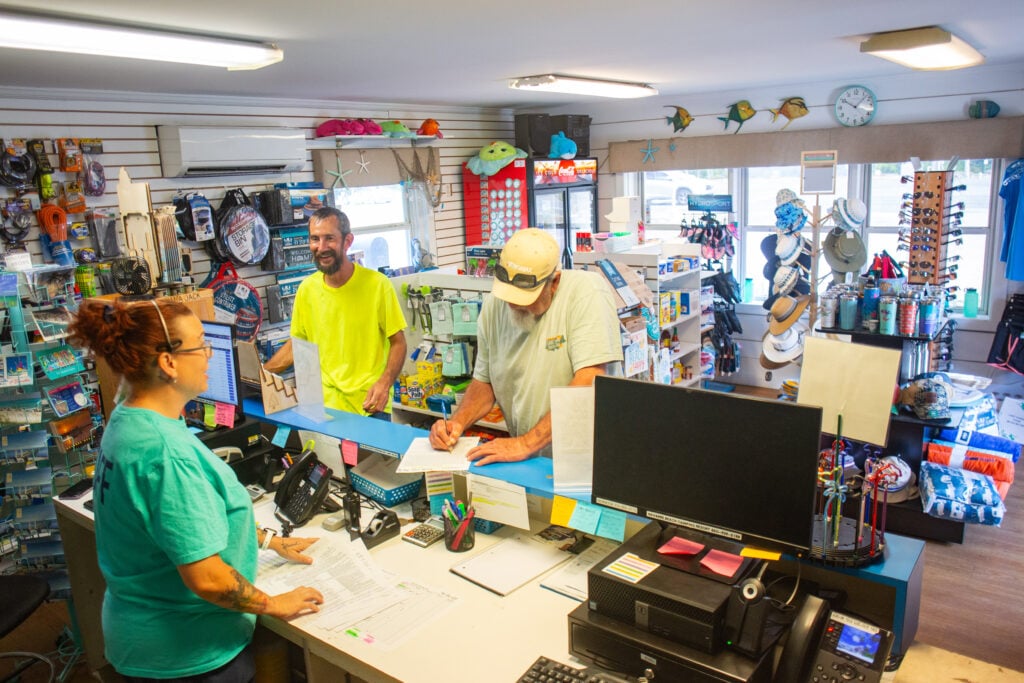 Three people interact at a store counter filled with papers and office supplies; shelves in the store display various merchandise and souvenirs.