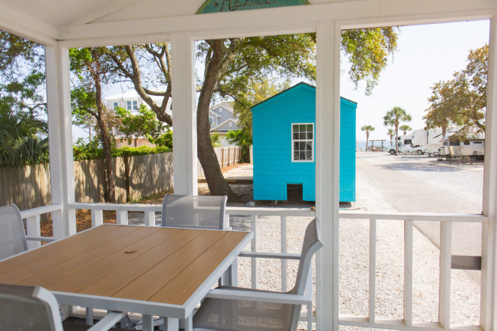 A view from a covered porch with a table and chairs, looking out toward a small bright blue building and a paved area with trees and houses in the background.