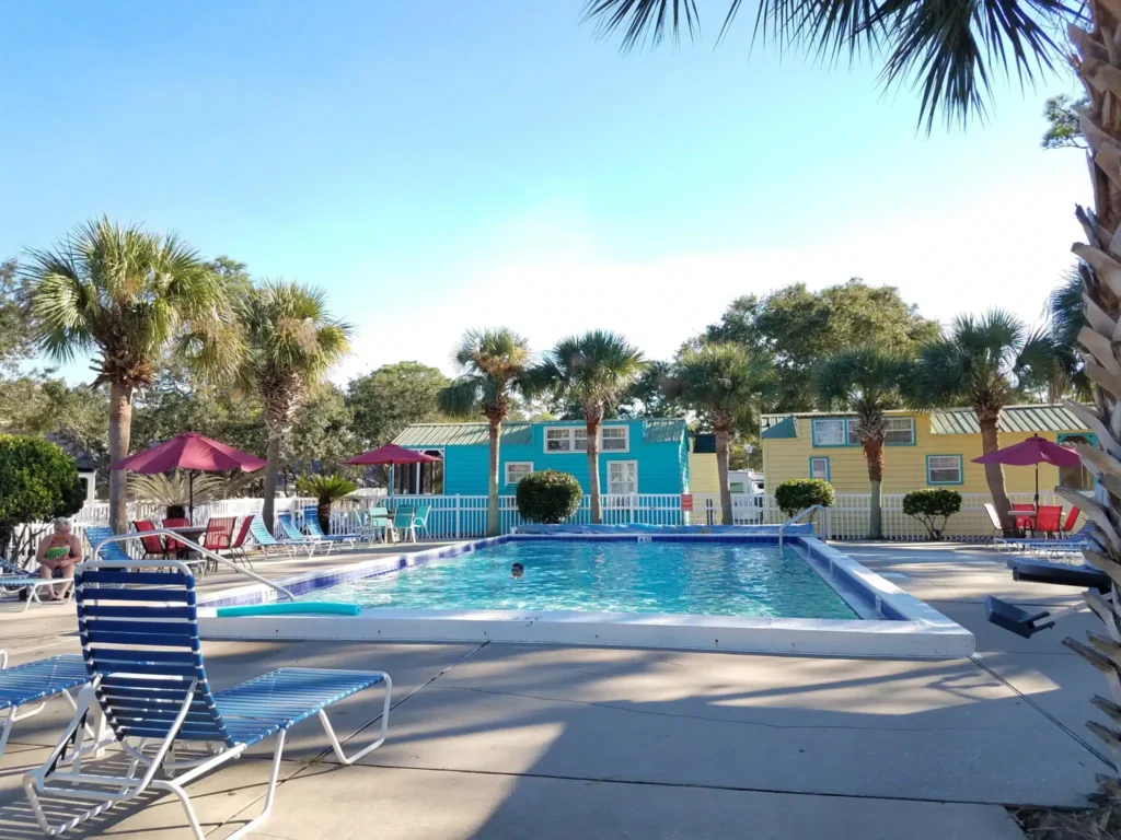 Outdoor swimming pool surrounded by lounge chairs, palm trees, and colorful buildings under a clear sky. A person is swimming in the pool.