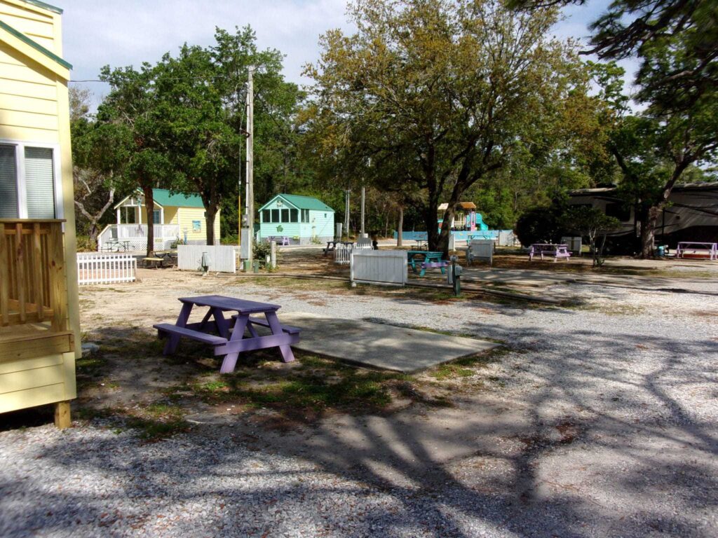 Gravel lot with purple picnic table, trees, small cabins, and benches; a camper is parked on the right side.