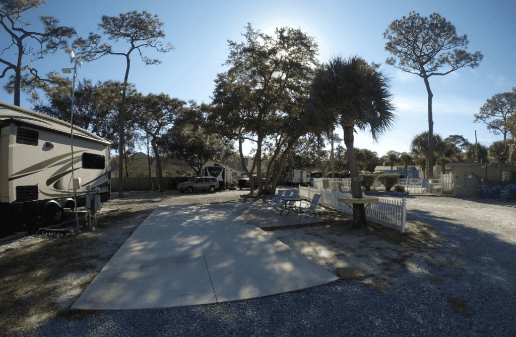 An empty RV parking spot with a picnic table and chairs, surrounded by trees and fencing in a sunlit campground.