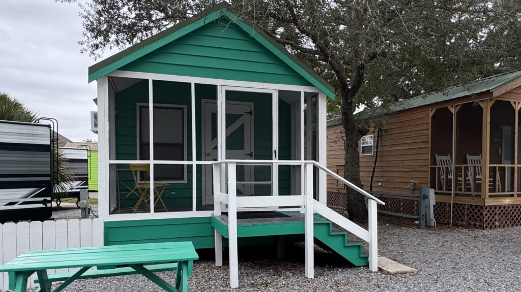 A small green cabin with a white screened porch, steps, and a yellow chair sits next to a brown cabin and a green picnic table on a gravel lot.