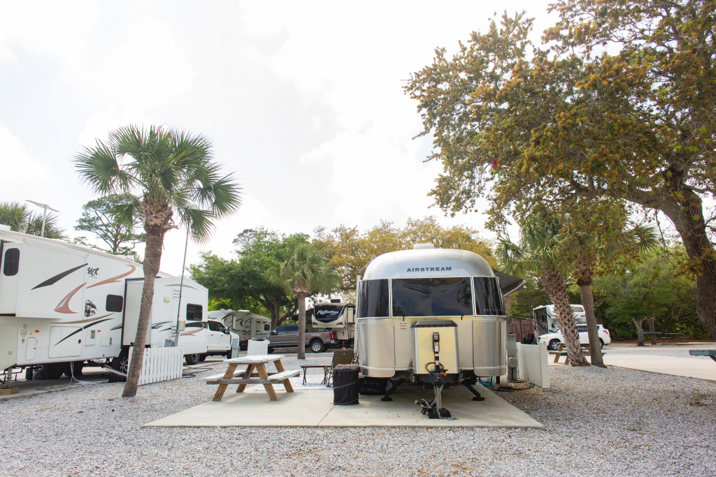A silver Airstream camper is parked at a gravel RV campsite with a picnic table and surrounded by trees and other RVs.