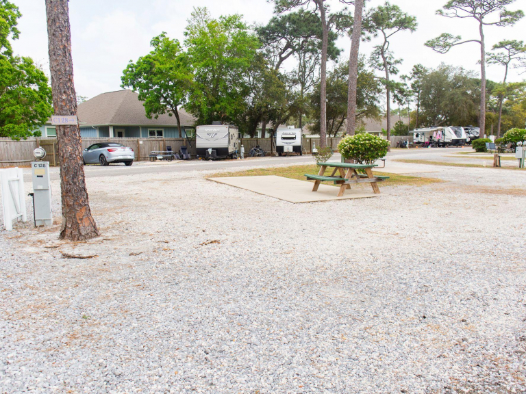 A gravel RV park with parked trailers, cars, a picnic table on a concrete pad, and trees with houses visible in the background.