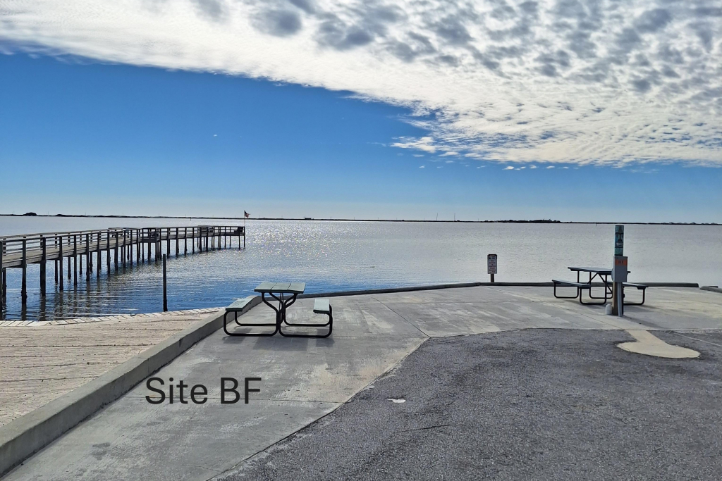 A boat launch site with two picnic tables overlooks calm water and a long pier under a partly cloudy sky. The area is labeled "Site BF.