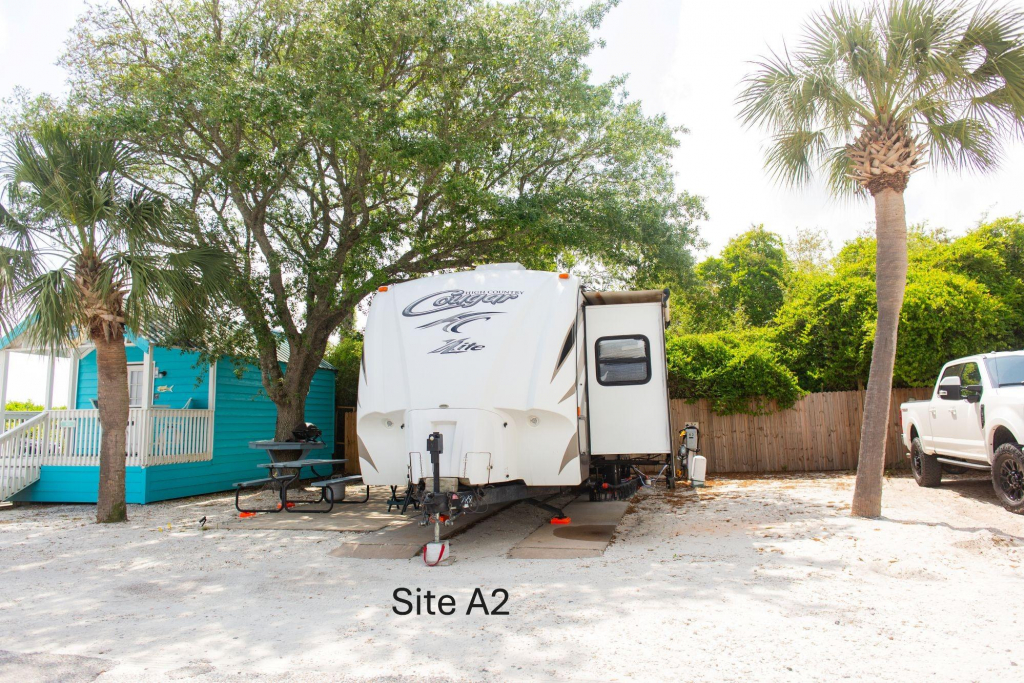 A travel trailer is parked at a sandy campsite labeled Site A2, with palm trees, a picnic table, a turquoise cabin, and a white truck nearby.