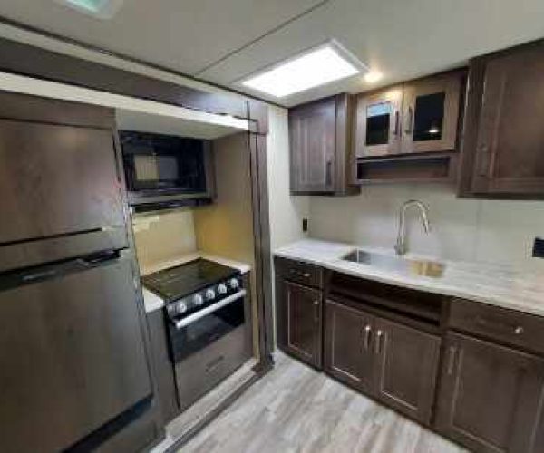 A modern kitchen featuring dark wood cabinets and stainless steel appliances. It includes a refrigerator, stove with oven, microwave, and a sink with overhead storage cabinets. The floor is covered with light-colored vinyl planks.