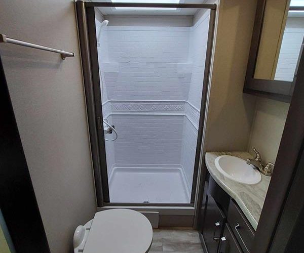 A small, modern bathroom with a glass-enclosed shower area, a white toilet, and a countertop with a sink and mirror. The walls are light-colored, and the flooring has a wood-like appearance. A towel rack is mounted on the wall near the shower.