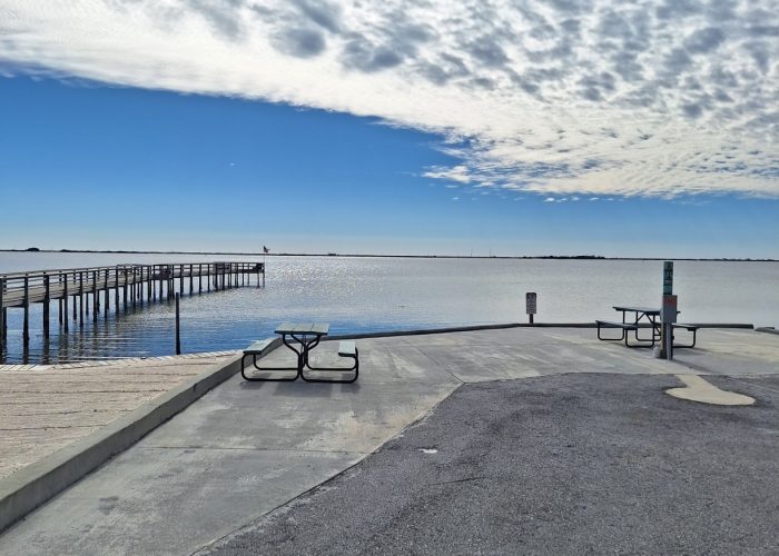 A paved area by the water with two picnic tables, a dock extending into the water, and a partly cloudy sky overhead.