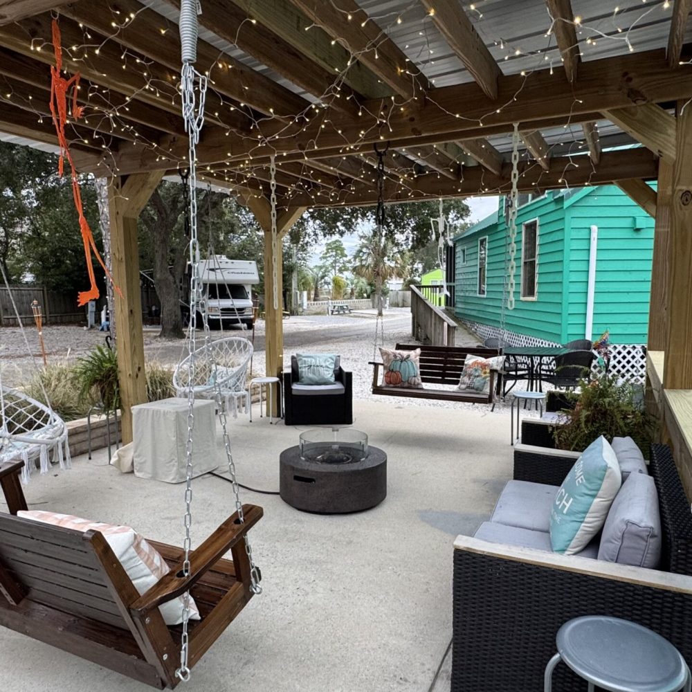 Outdoor patio under a wooden pergola with string lights, cushioned chairs, a wooden swing, a fire pit, and a turquoise building in the background.
