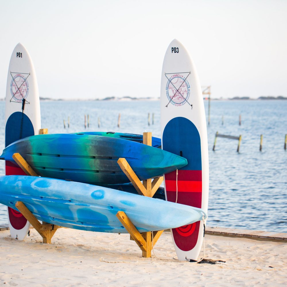 Two surfboards are upright in the sand behind two stacked kayaks on a wooden rack by the edge of a calm body of water.