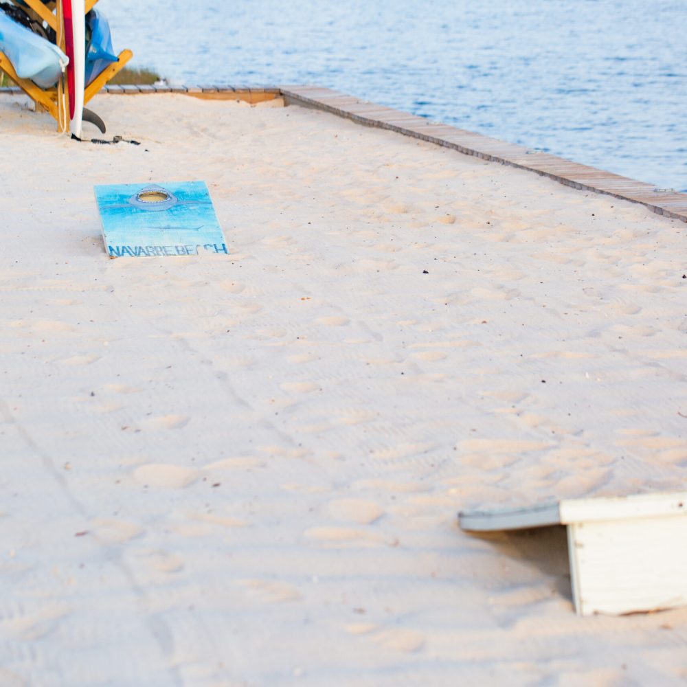 A cornhole game set is placed on sandy ground near a wooden deck by the water, with paddles stored nearby.
