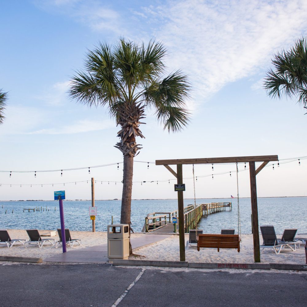 A sandy beach area with palm trees, lounge chairs, string lights, and a wooden pier extending into calm blue water under a partly cloudy sky.