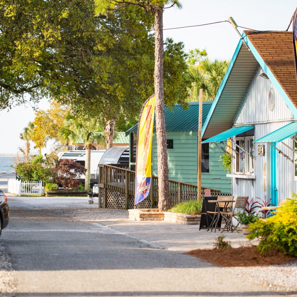 A small street lined with cottages, a car parked on the left, colorful flags advertising wine and beer, and trees with water visible in the background.