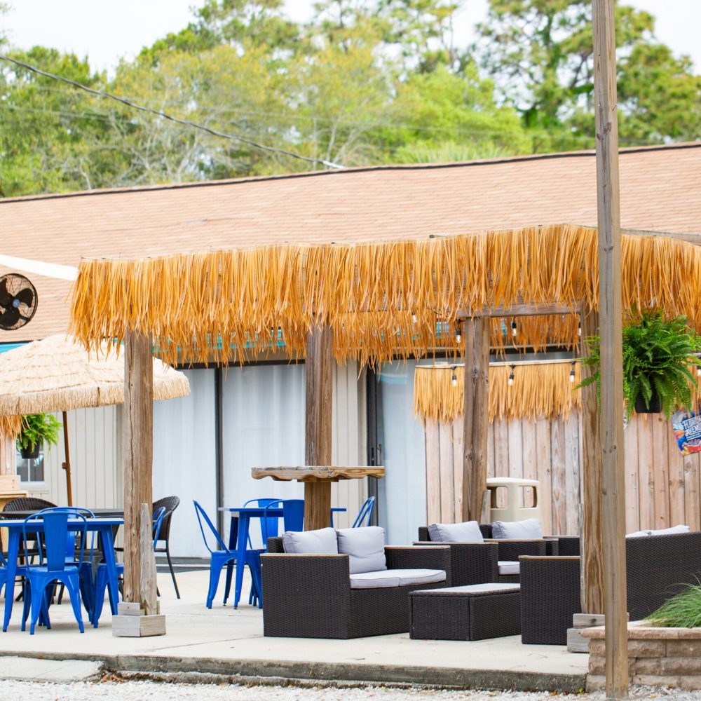 Outdoor seating area with wicker furniture, blue chairs, tables, and thatched-roof pergolas, surrounded by a wooden fence and greenery.