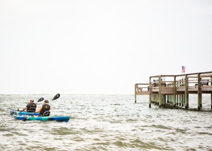 Two people paddle a blue kayak near a wooden pier over the water, with an American flag visible on the pier.