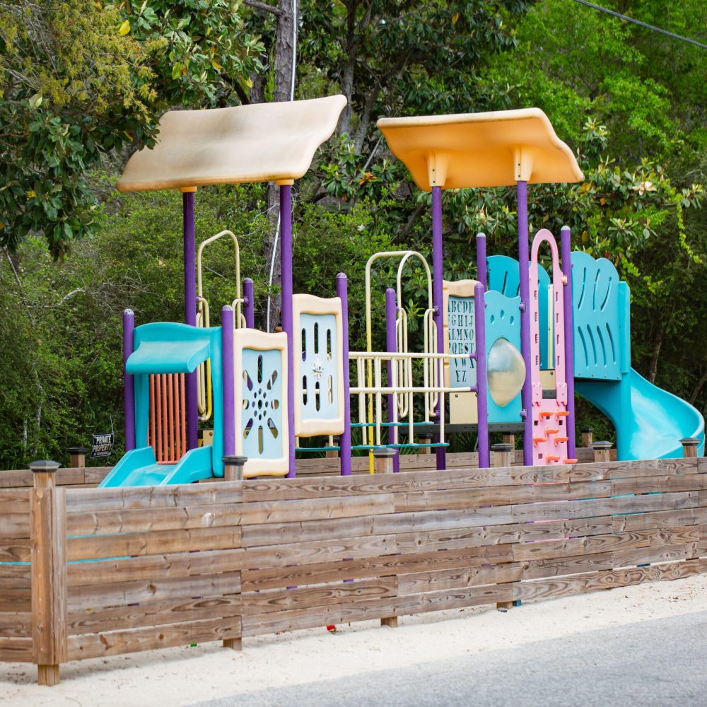 A colorful outdoor playground with slides and climbing structures is enclosed by a wooden fence, set against a backdrop of trees and greenery.