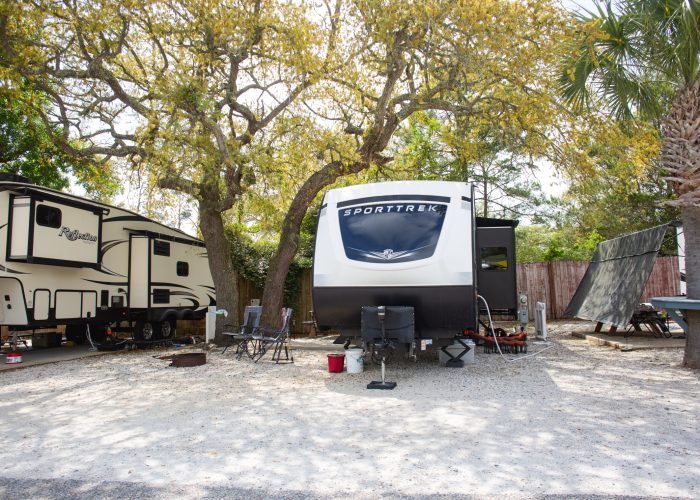 Two RVs are parked side by side on gravel under large trees, with lawn chairs, a cooler, and other camping items set up outside.