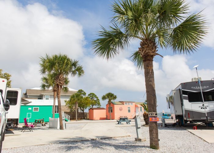 RV park with palm trees, colorful tiny homes, gravel lots, and picnic tables under a partly cloudy sky.