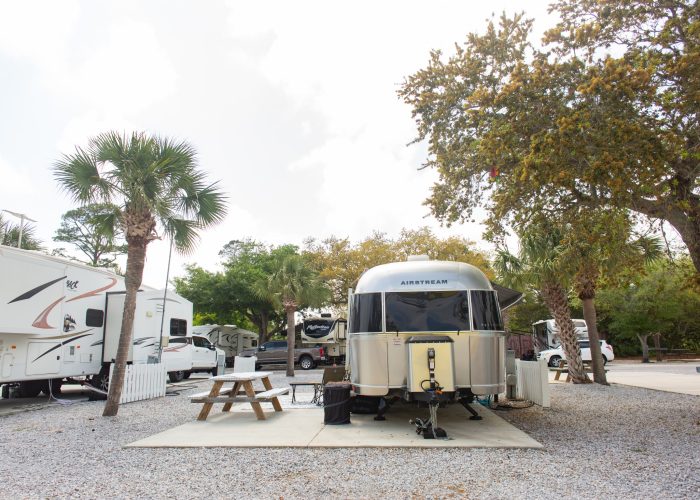 A silver Airstream trailer is parked on a gravel lot with a picnic table nearby, surrounded by trees and other RVs at a campground.