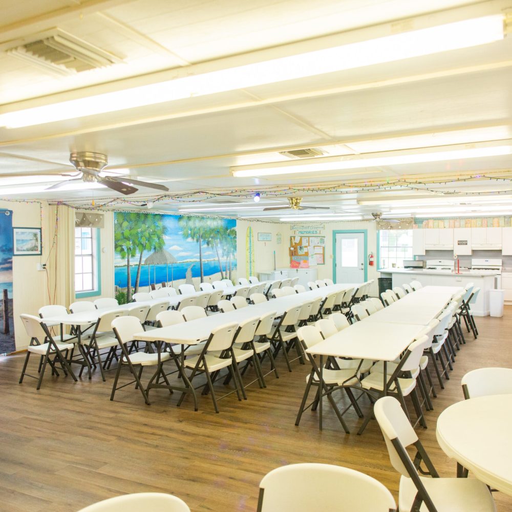 A brightly lit room with long tables and white folding chairs arranged in rows, tropical beach murals on the walls, a kitchen area in the back, and wooden floors.