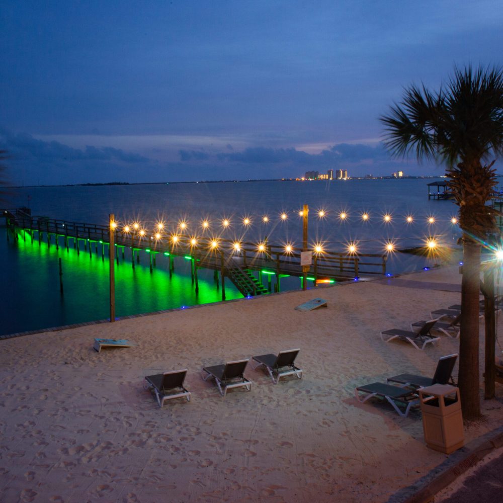 Sandy beach at dusk with lounge chairs, palm trees, and a lit pier extending over calm water, illuminated by green and white lights.