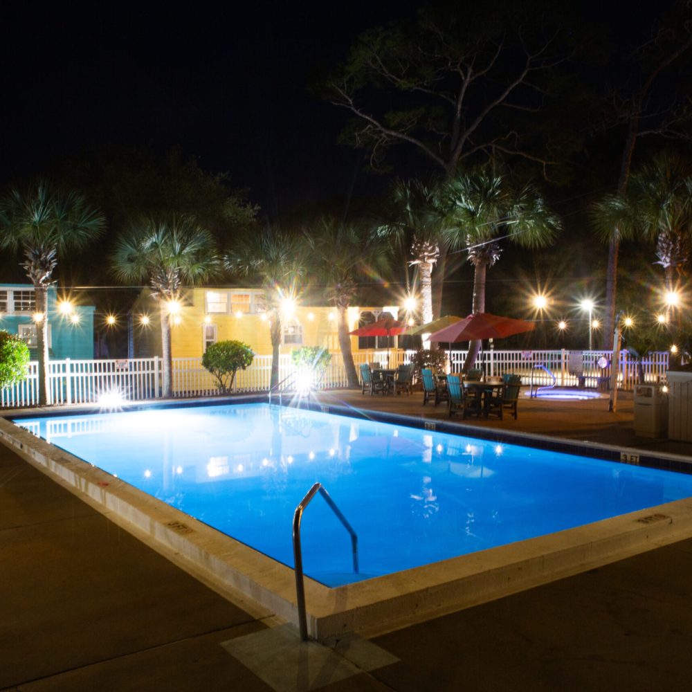 A rectangular outdoor swimming pool is illuminated at night, surrounded by lounge chairs, palm trees, string lights, and patio tables with umbrellas.
