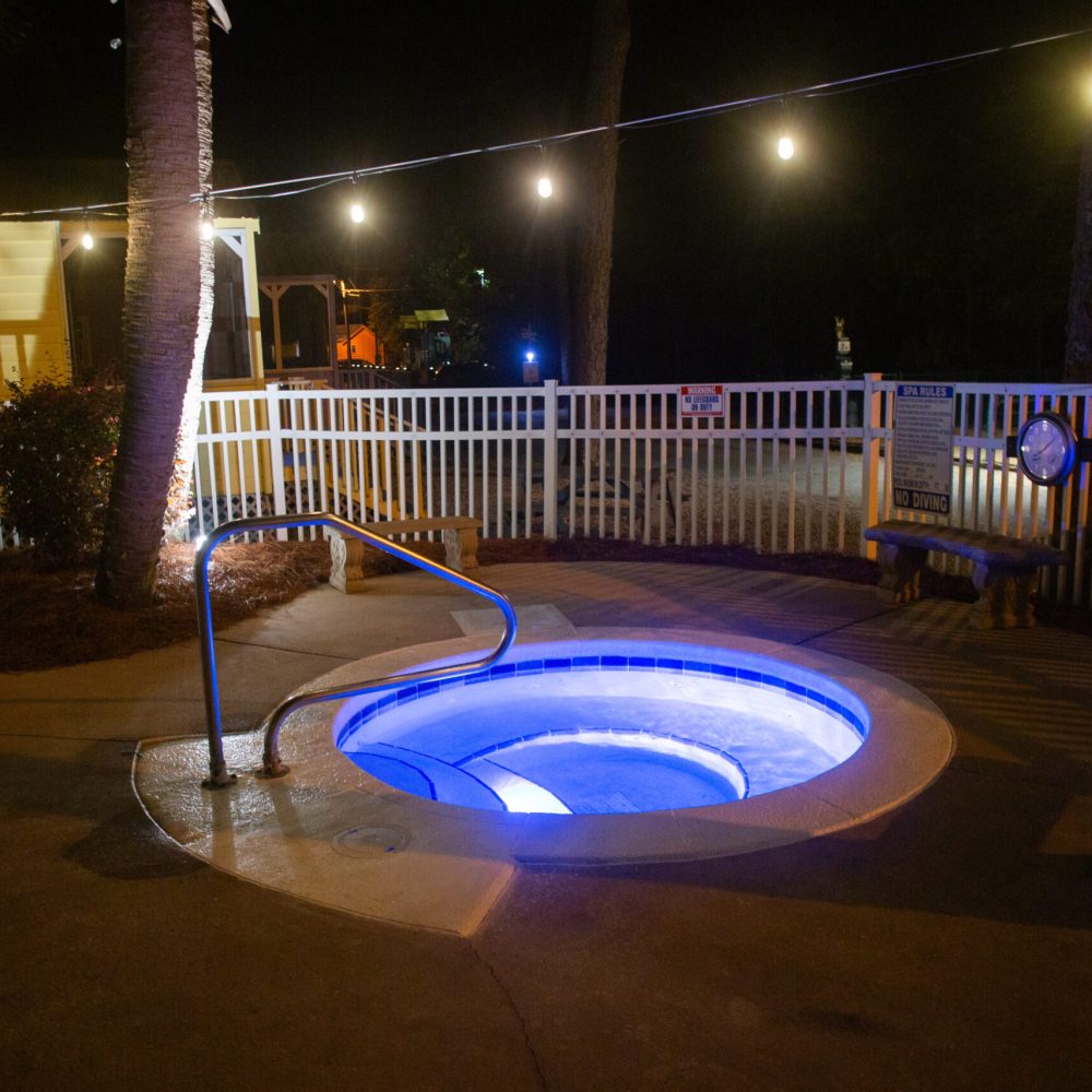 A small, circular hot tub with blue lights is surrounded by a white fence and outdoor string lights at night.