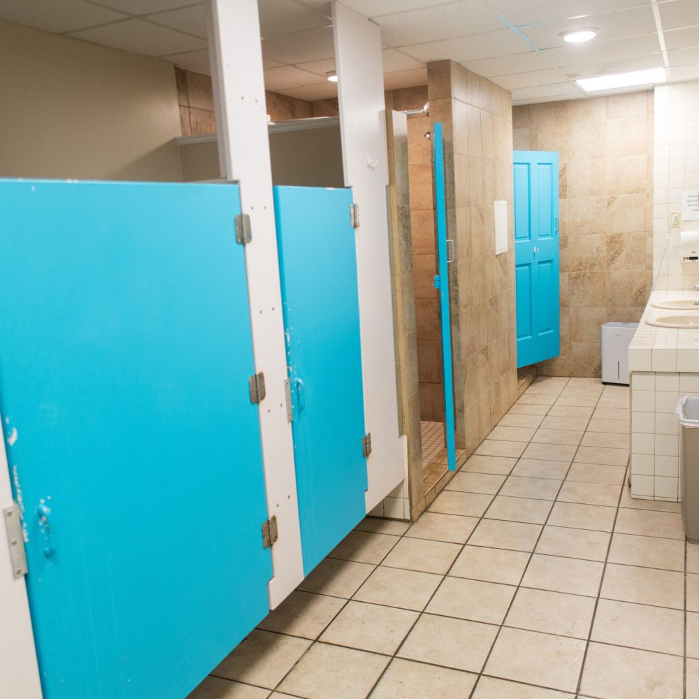 A public restroom with blue stall doors, beige tiled floor, sinks with a mirror, a trash can, and overhead fluorescent lighting.