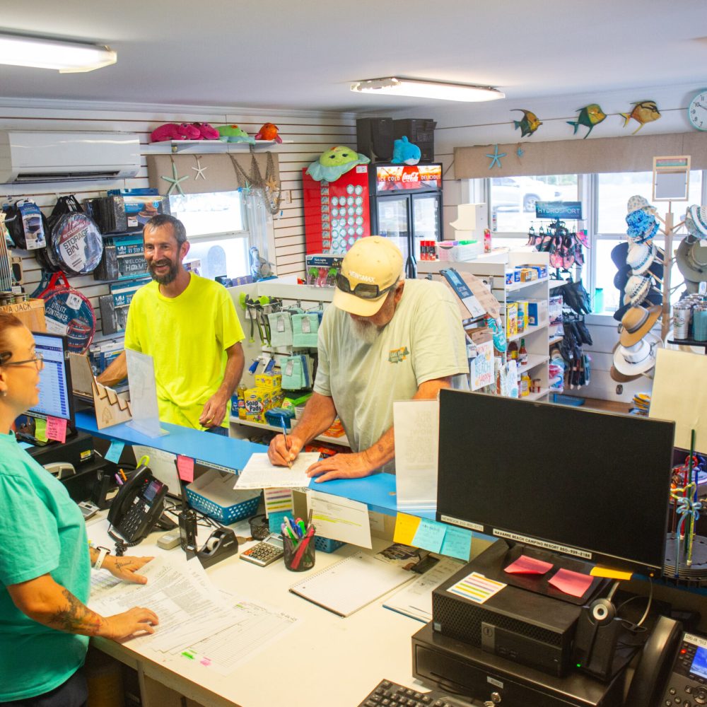 Three people interact at the counter of a small retail store filled with various merchandise and supplies, including hats, sunglasses, and snacks.