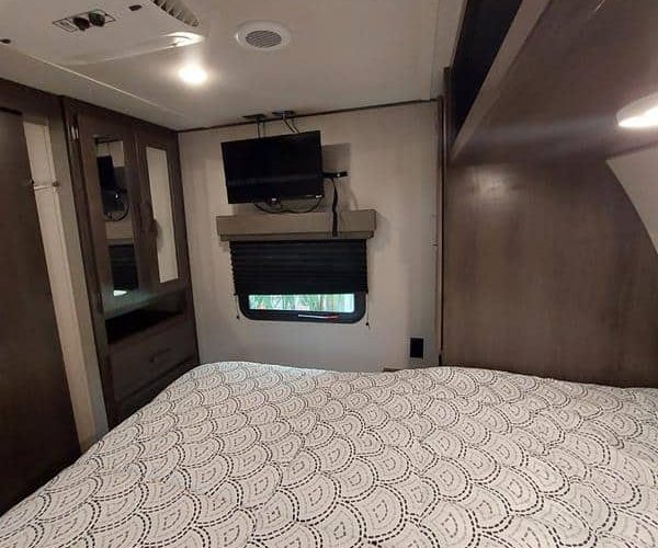 Interior of a cozy RV bedroom with a patterned bedspread on a bed. The room features a small wall-mounted TV above a window with a black roller blind. The walls and furniture are in shades of brown, with a door and a set of mirrored cabinets on the left.