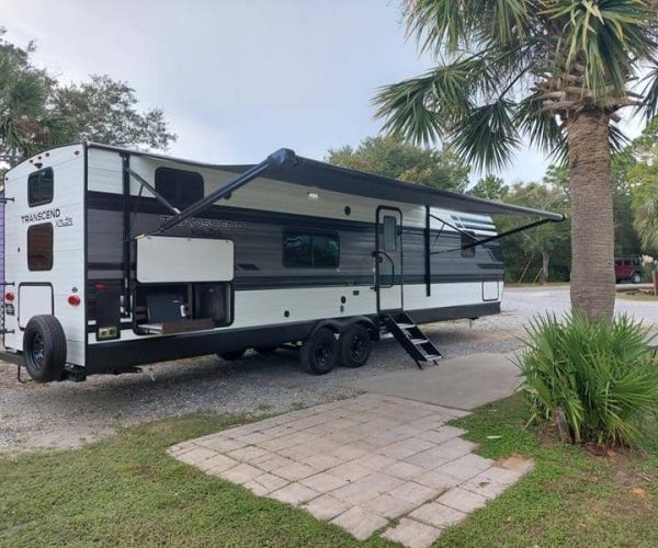 A white and black travel trailer with an extended awning is parked in a campground next to a palm tree. The trailer has multiple wheels, slide-out sections, and an outdoor kitchen area. There are other RVs and trees visible in the background.
